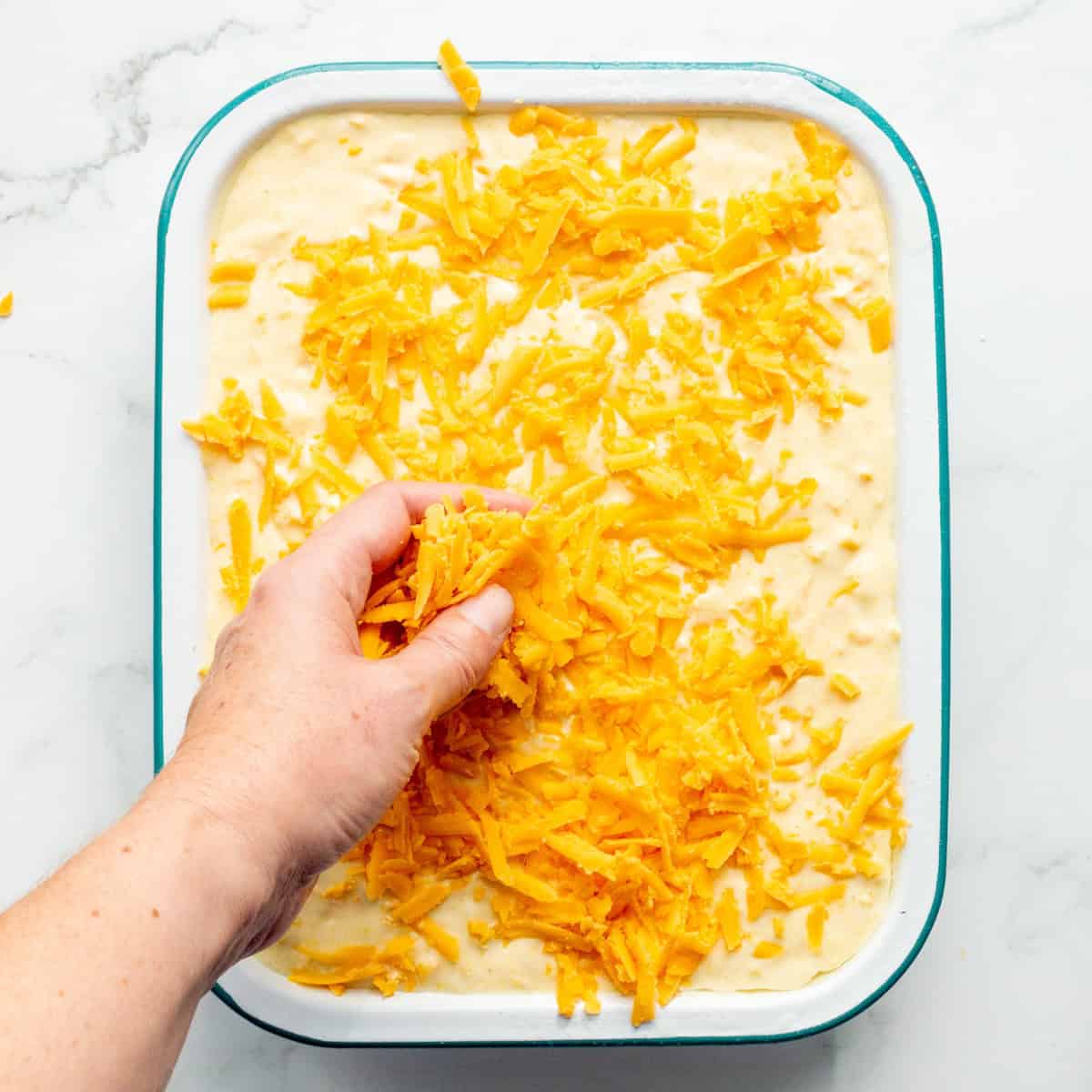 Sprinkling shredded Cheddar cheese on top of the corn casserole batter.