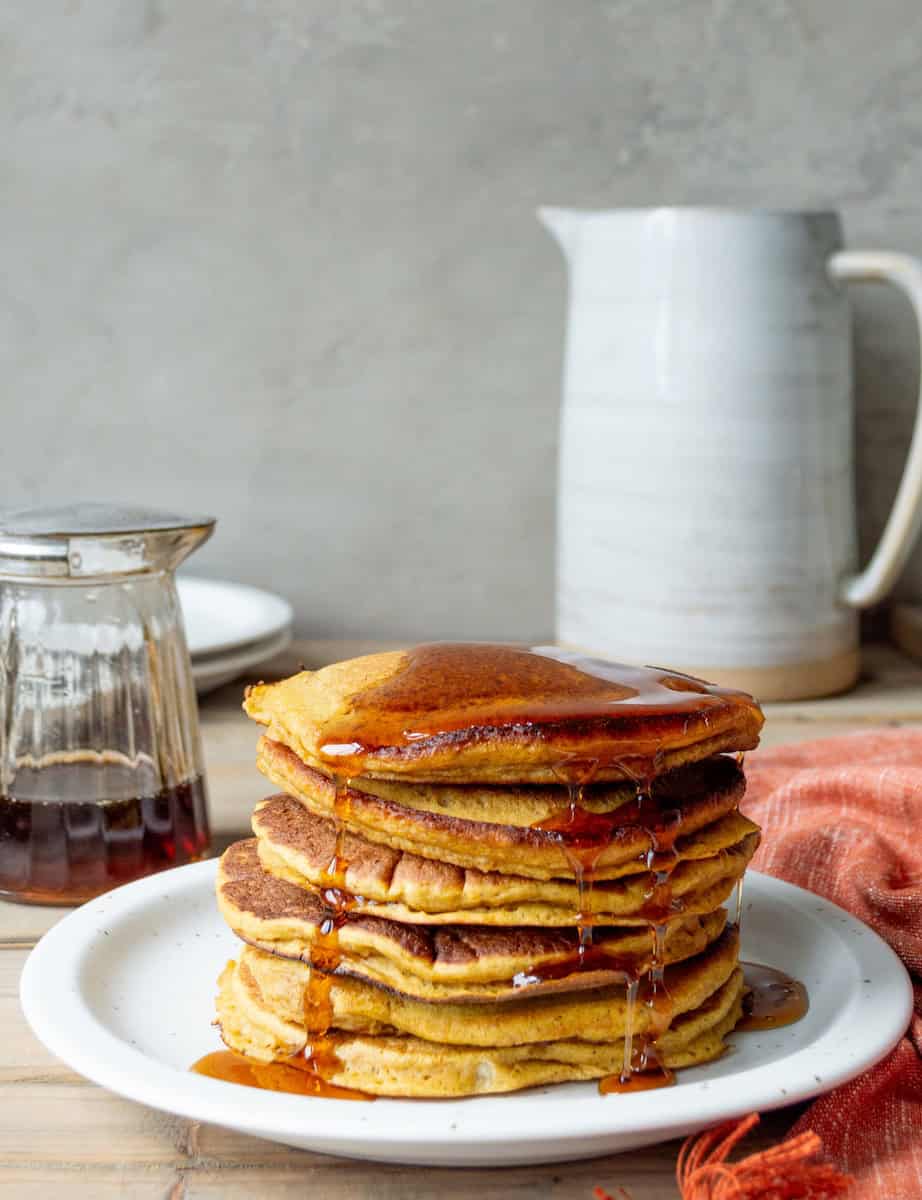 Trader Joe's pumpkin pancakes on a white plate with an orange cloth, syrup pitcher, white pitcher, and stack on plates on the side.