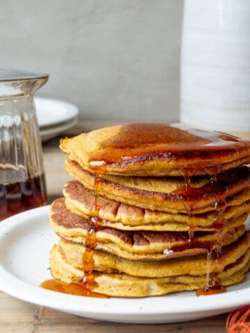 Trader Joe's pumpkin pancakes on a white plate with an orange cloth, syrup pitcher, white pitcher, and stack on plates on the side.