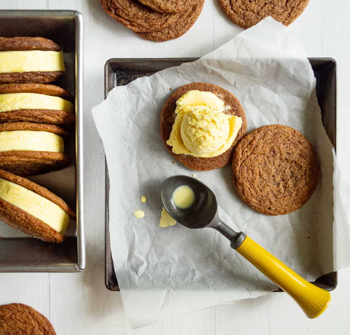 Ginger Molasses Cookies being made into Lemon Custard Ginger Molasses Ice Cream cookie sandwiches in a metal tin lined with parchment paper and a yellow handled ice cream scoop.