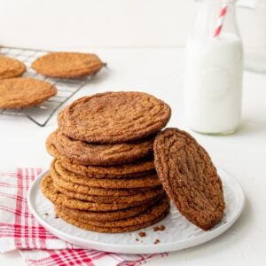 A white plate with a stack of chewy gingerbread cookies and a red checked napkin plus a wire rack of cookies and bottle of milk with a red and white striped straw in the background.