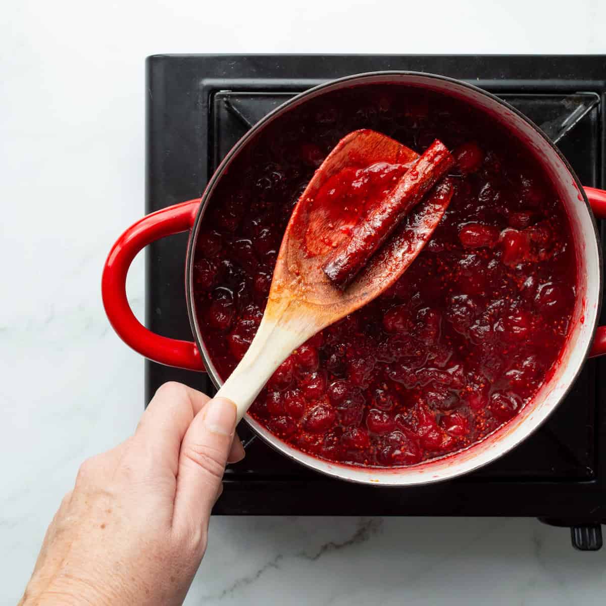 Removing a cinnamon stick from cranberry sauce in a red handled pot.