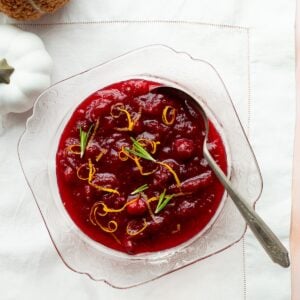 Orange cranberry sauce in a pink glass bowl on a white linen with a white pumpkin and fuzzy orange pumpkin on the side.