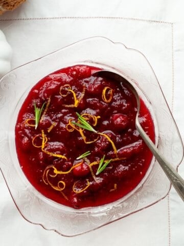 Orange cranberry sauce in a pink glass bowl on a white linen with a white pumpkin and fuzzy orange pumpkin on the side.