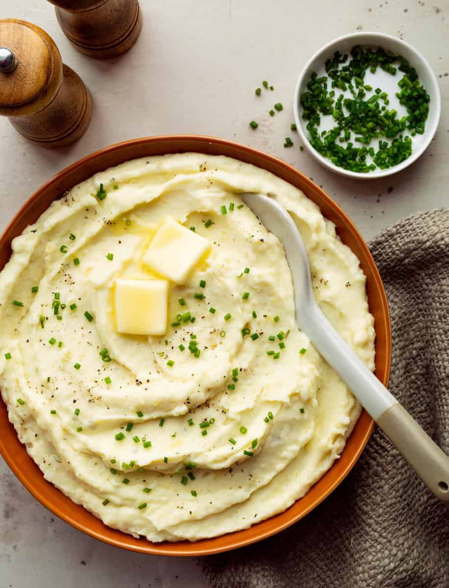An orange bowl of creamy boursin mashed potatoes topped with two butter pats and chopped chives plus a white bowl of chives, a brown cloth, and wooden salt and pepper shakers off to the side.