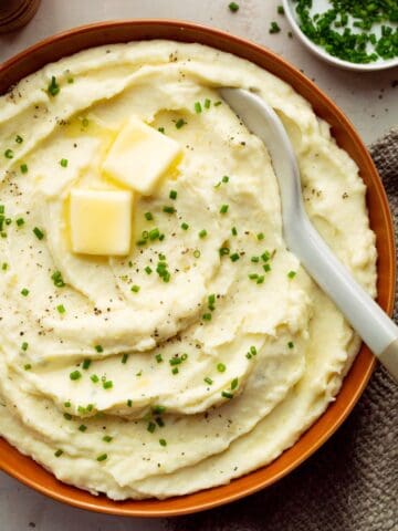An orange bowl of creamy boursin mashed potatoes topped with two butter pats and chopped chives plus a white bowl of chives, a brown cloth, and wooden salt and pepper shakers off to the side.