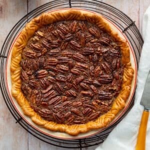 Bourbon pecan pie in a red rimmed ceramic pie dish on a round wire rack with a white cloth and yellow handled pie server.