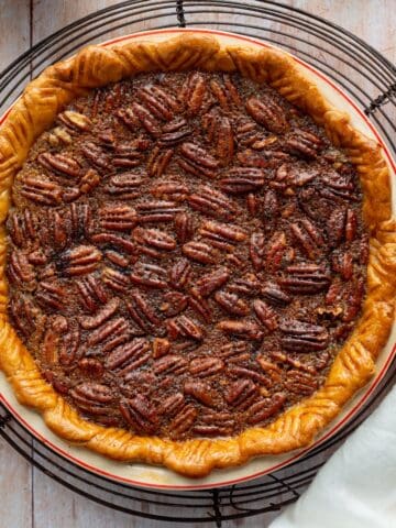 Bourbon pecan pie in a red rimmed ceramic pie dish on a round wire rack with a white cloth and yellow handled pie server.