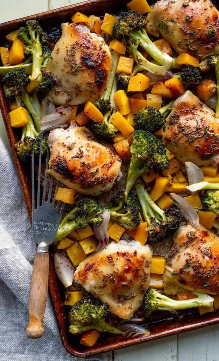 Sheet pan honey mustard chicken thighs and veggies with a vintage serving spatula and a beige cloth.