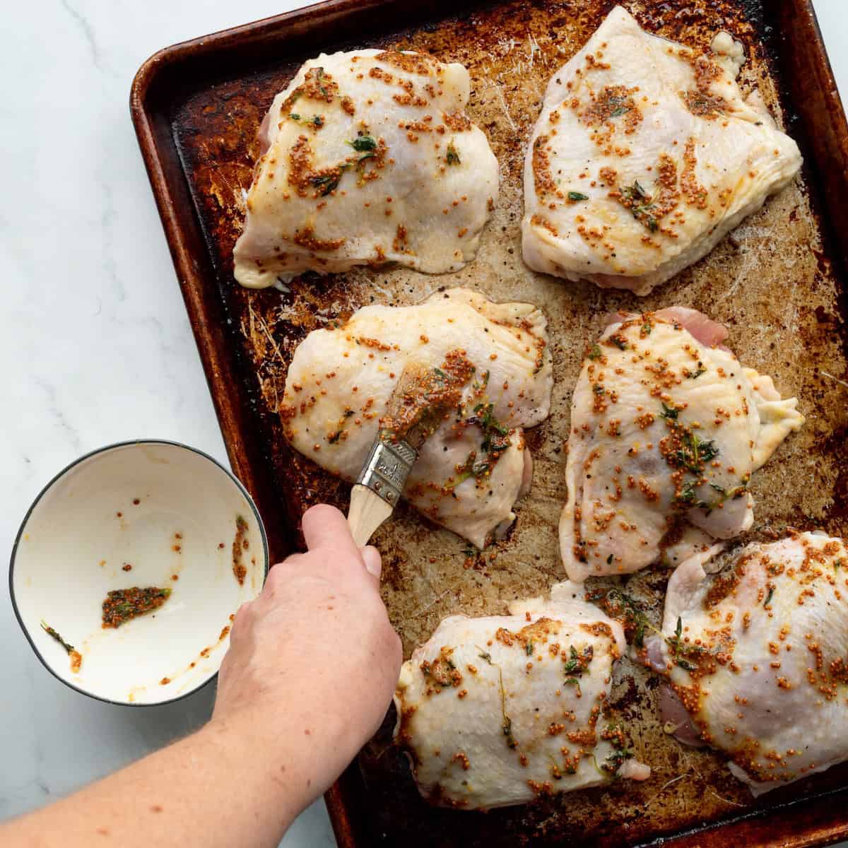 Brushing a honey, whole grain mustard, and thyme mixture onto chicken thighs on a rimmed baking sheet.