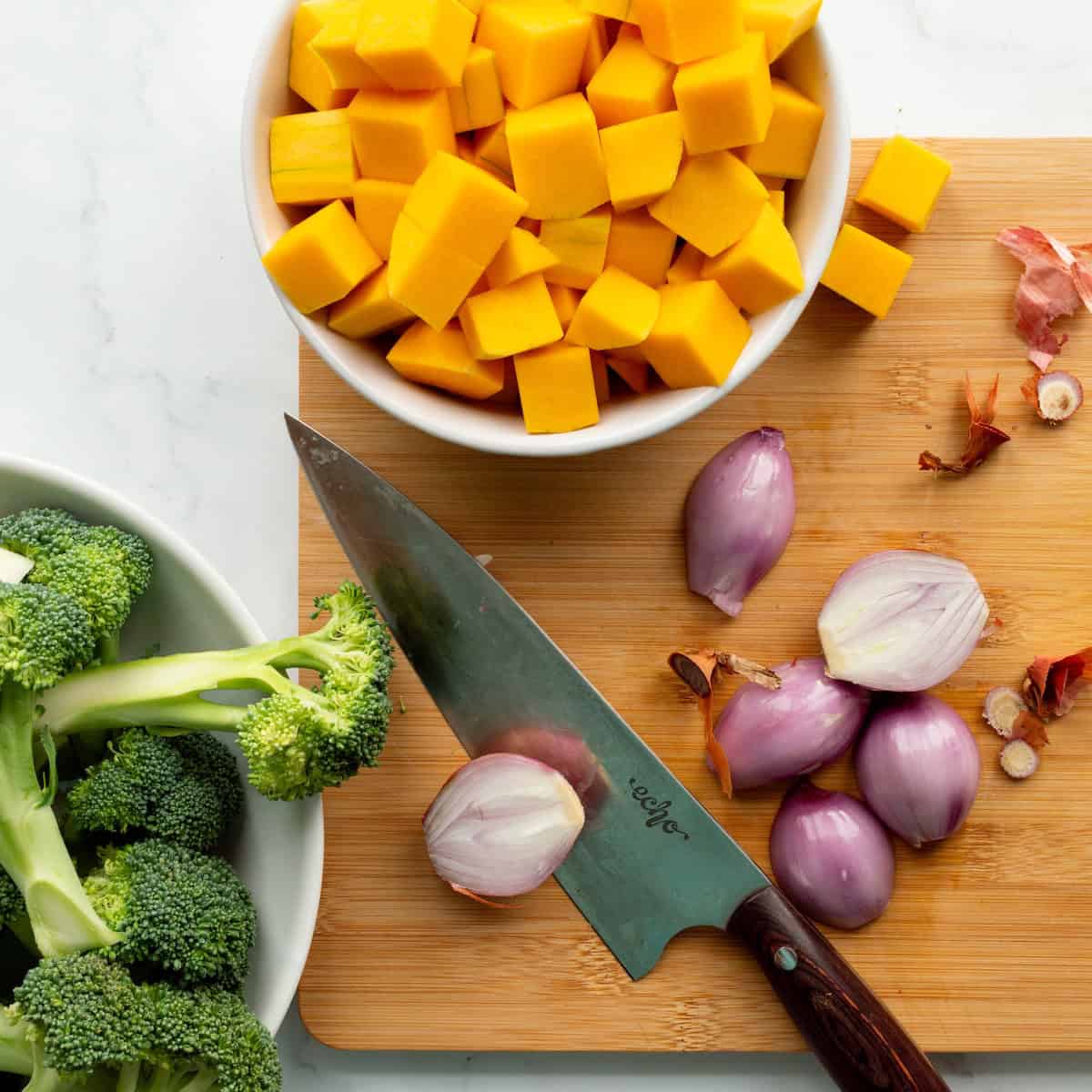 Cubed butternut squash in a white bowl, a white bowl of broccoli spears, and halved shallots on a wooden cutting board with a chef's knife.