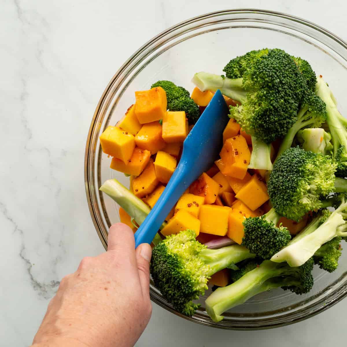 Stirring together the broccoli, shallots, and butternut squash with a honey mustard vinaigrette in a glass bowl with a blue spatula.