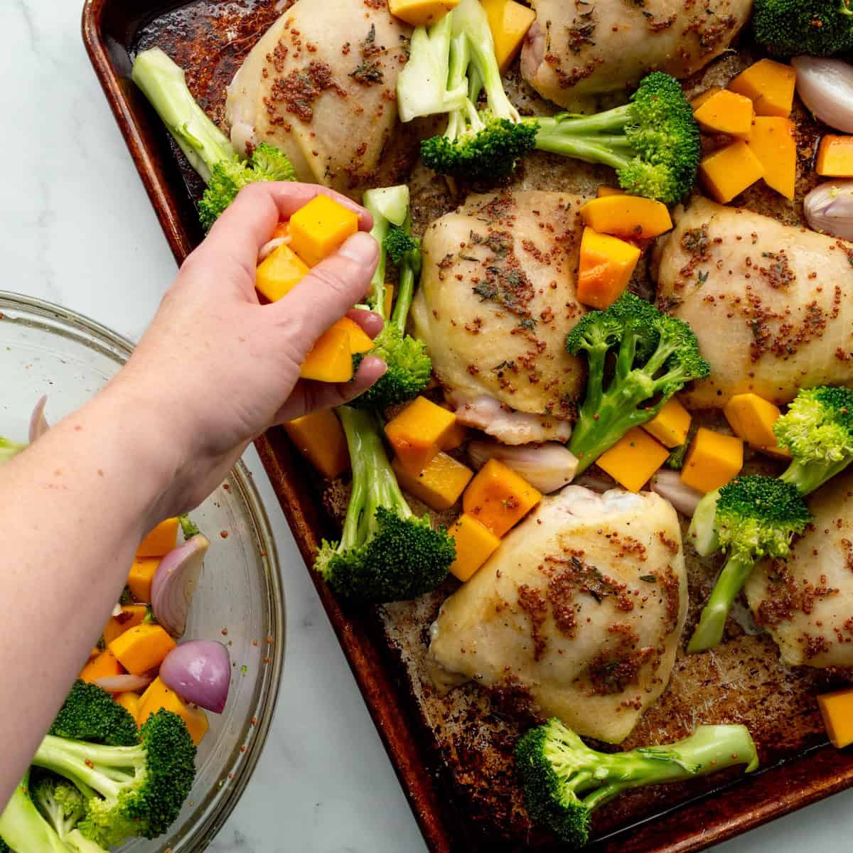 Adding butternut squash, shallots, and broccoli, to a sheet pan of honey mustard chicken thighs.
