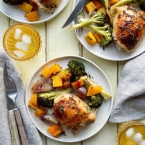 Three cream colored plates of sheet pan chicken thighs and vegetables with a yellow glass of ice water, wooden handled utensils, and beige napkins.