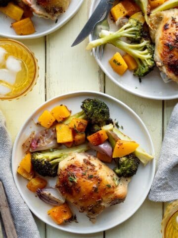 Three cream colored plates of sheet pan chicken thighs and vegetables with a yellow glass of ice water, wooden handled utensils, and beige napkins.