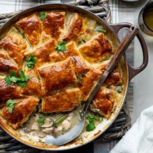 Puff pastry turkey pot pie in a dark brown skillet on a wicker trivet and plaid cloth with a plate of pot pie, dish of fresh herbs, and dark glasses on the side.