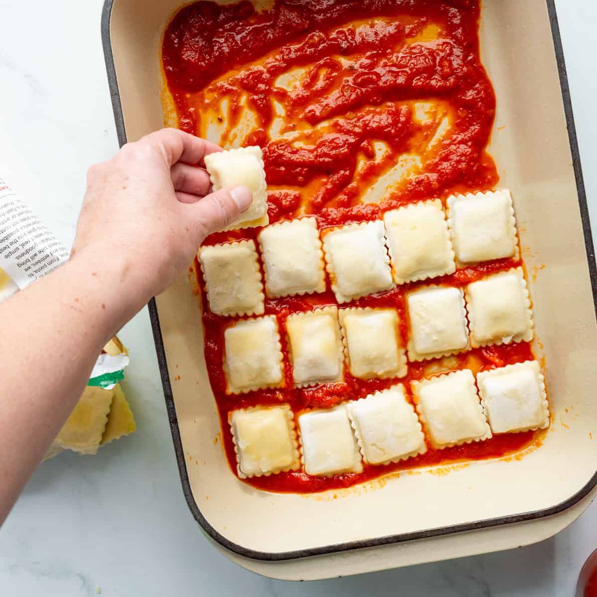 Arranging a single layer of frozen cheese ravioli on red sauce in a casserole dish.
