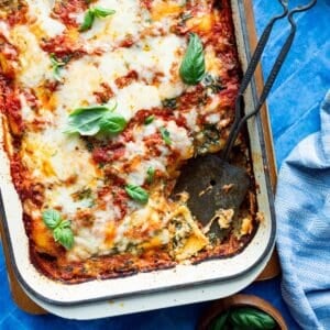 A casserole dish of Lazy Frozen Cheese Ravioli Lasagna on a wooden board with a metal scooper and wooden bowl of fresh basil.