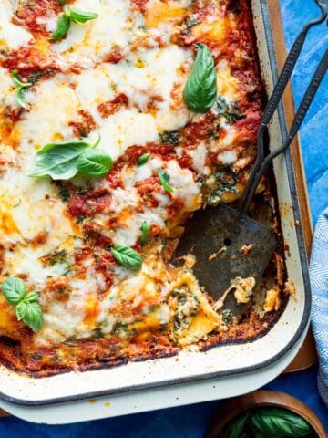 A casserole dish of Lazy Frozen Cheese Ravioli Lasagna on a wooden board with a metal scooper and wooden bowl of fresh basil.