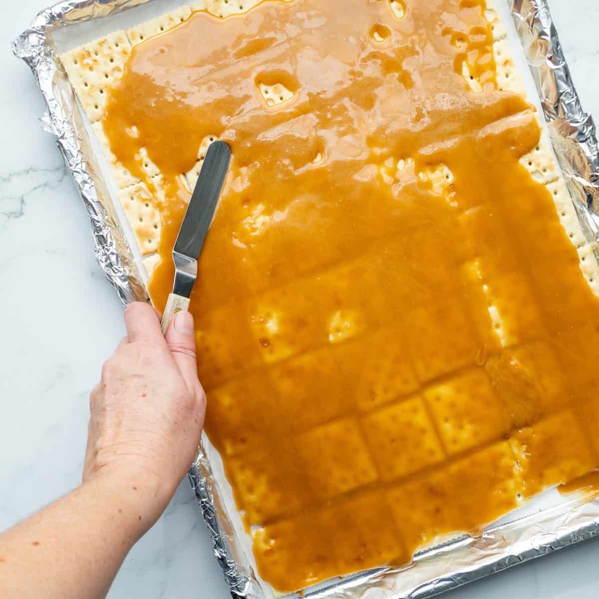 Spreading toffee onto a layer of saltine crackers in an aluminum foil lined baking sheet.