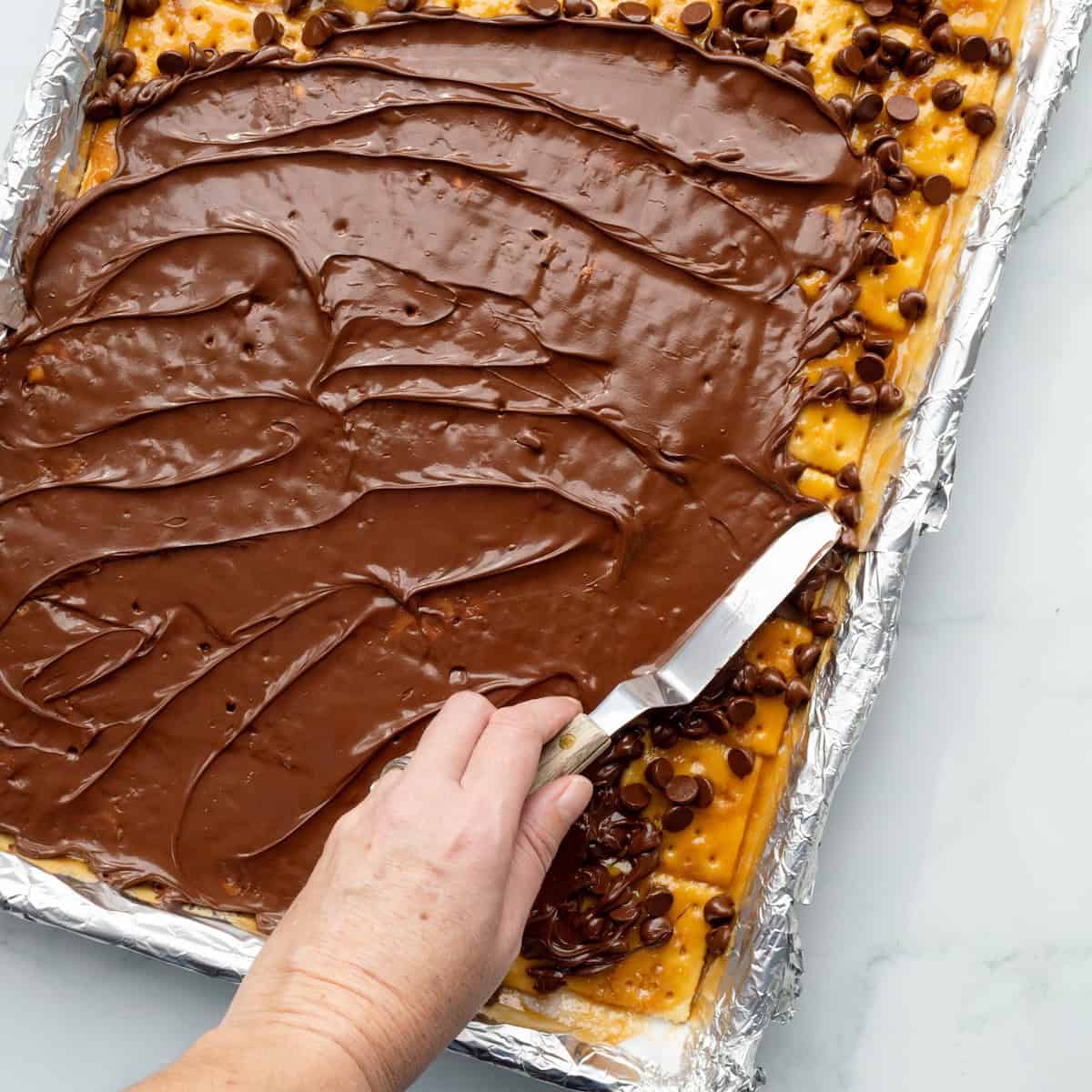 Spreading melted chocolate chips on top of toffee covered saltine crackers in an aluminum foil lined baking pan.