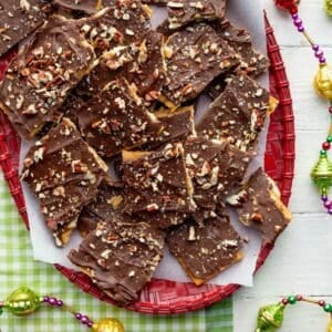 Red basket weave platter topped with chocolate saltine cracker toffee on a green checked cloth surrounded by Christmas garland.