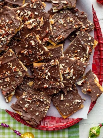 Red basket weave platter topped with chocolate saltine cracker toffee on a green checked cloth surrounded by Christmas garland.