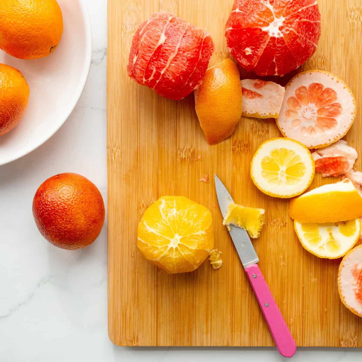 Cutting the peel from grapefruit and oranges with a pink paring knife on a wooden cutting board.