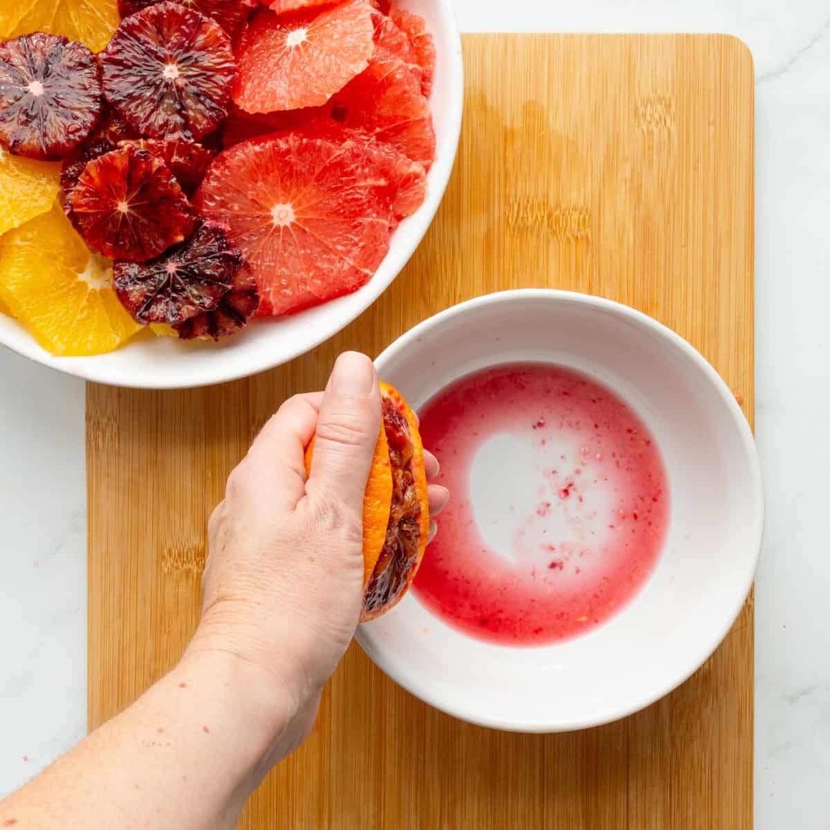 Squeezing blood orange juice into a white bowl with a bowl of sliced citrus to the side.