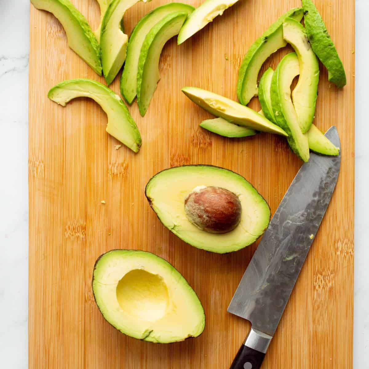 Halved, pitted, and sliced avocado on a wooden cutting board with a chef's knife.