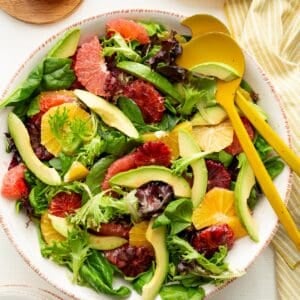 Winter citrus and avocado salad in a large pottery bowl with yellow salad servers, surrounded by salad plates, a pitcher of dressing, and a yellow striped towel.