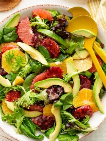 Winter citrus and avocado salad in a large pottery bowl with yellow salad servers, surrounded by salad plates, a pitcher of dressing, and a yellow striped towel.