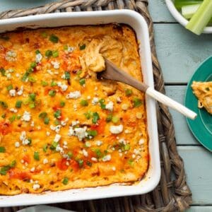 A white square dish of cottage cheese buffalo chicken dip on a basket weave trivet along with small plates, celery sticks, and chips on the side.
