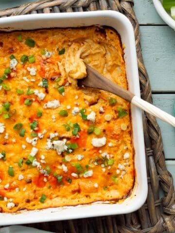 A white square dish of cottage cheese buffalo chicken dip on a basket weave trivet along with small plates, celery sticks, and chips on the side.