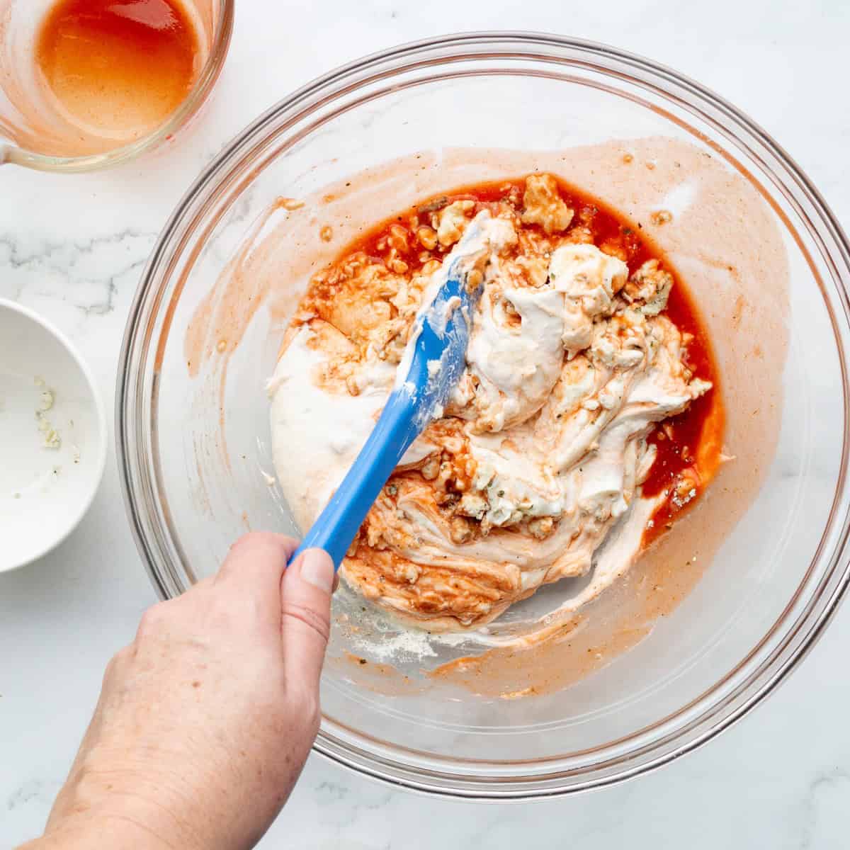 Stirring together the ingredients for high protein buffalo dip in a glass bowl with a blue rubber spatula.
