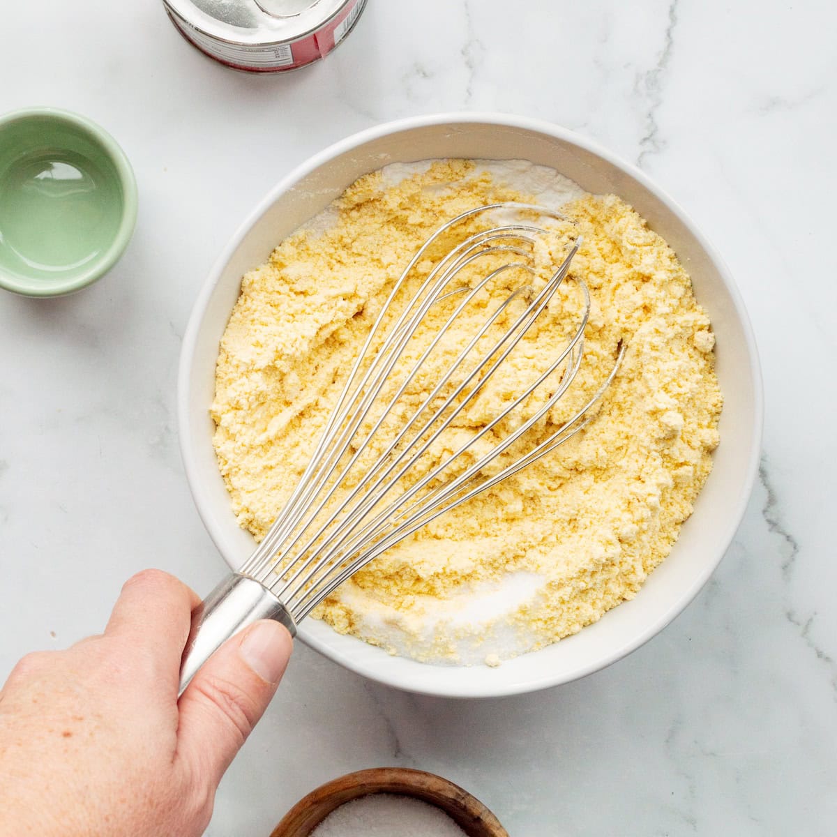 Whisking together yellow cornmeal, baking soda, baking powder, sugar, and salt in a white bowl.