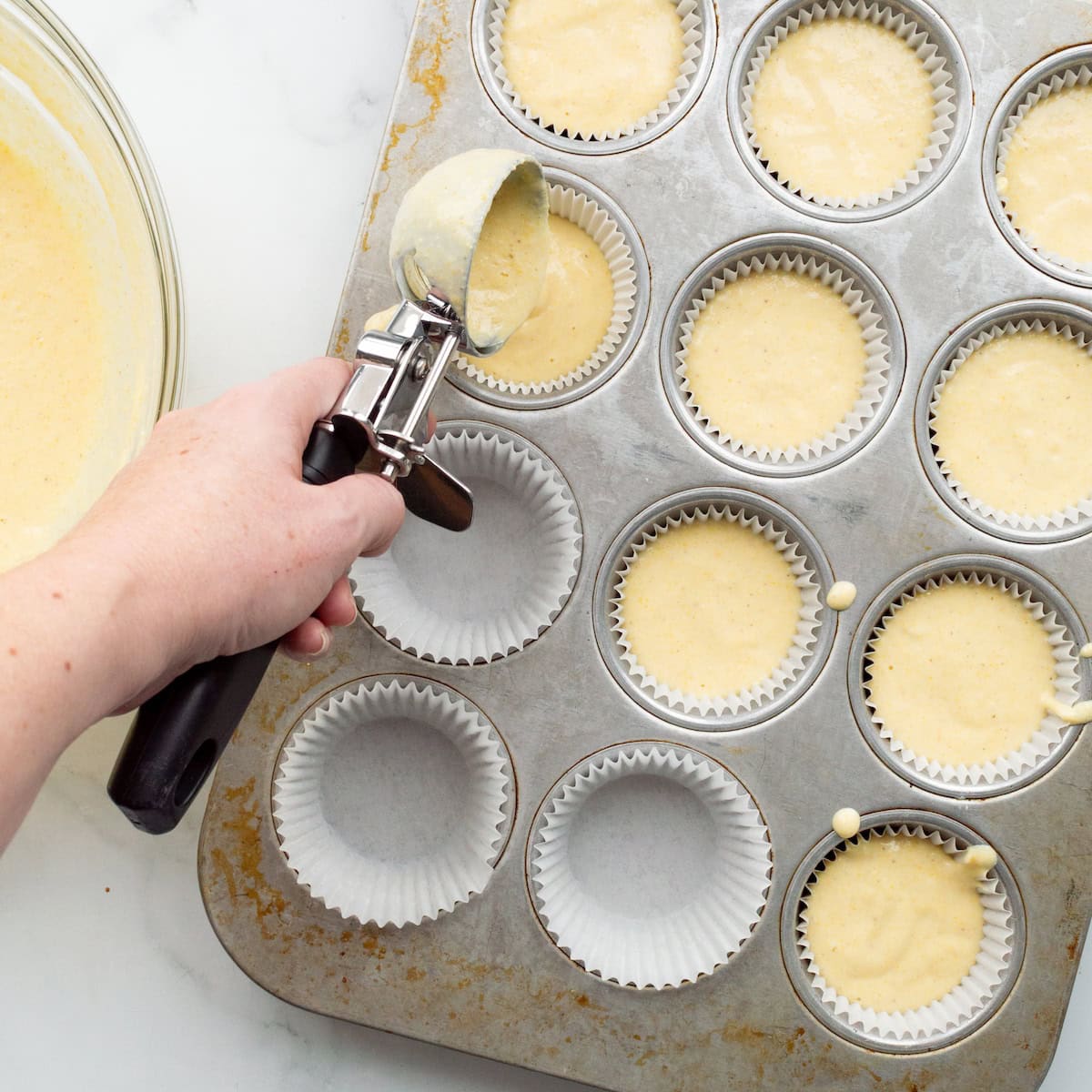 Scooping buttermilk cornbread batter into paper liners in a muffin pan with an ice cream scoop.