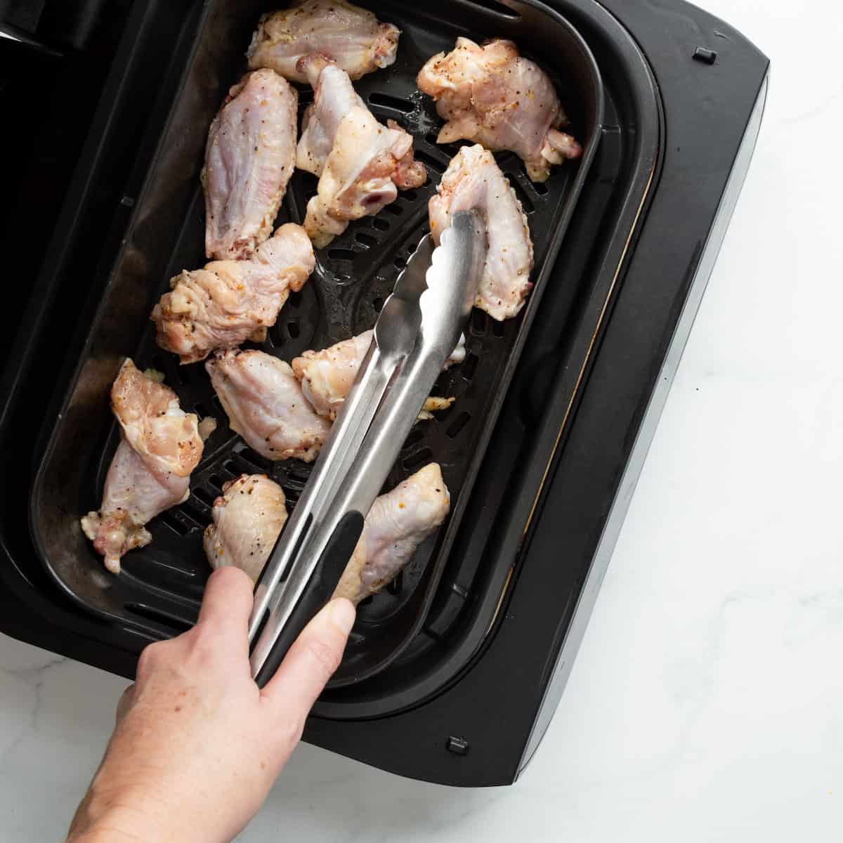 Adding seasoning raw chicken wings to an air fryer with metal tongs.