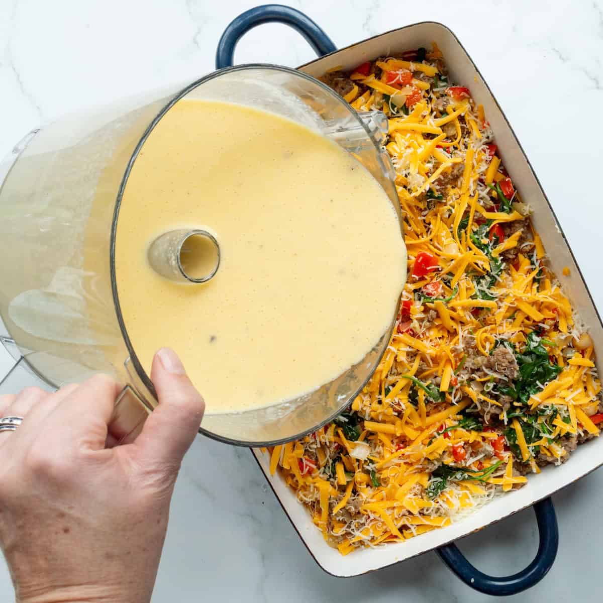 Pouring the cottage cheese and egg mixture out of a food processor over a casserole dish filled with shredded cheese, sausage, and vegetables.