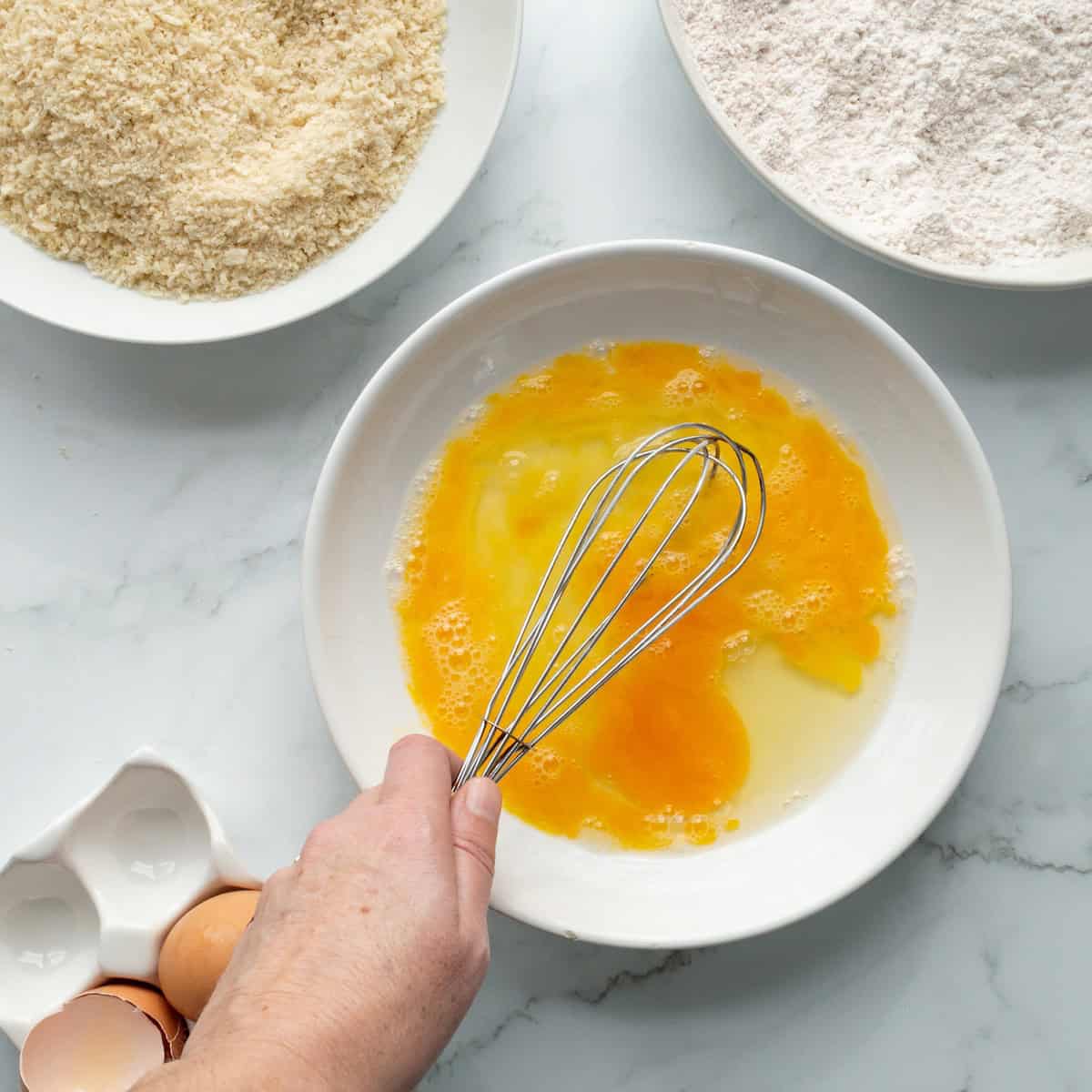 Whisking eggs in a shallow white bowl with flour and panko in separate shallow bowls to the side.