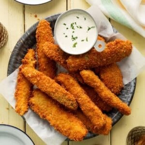 Fried pickle spears, also known as frickles, in a metal pie pan along with a cup of homemade ranch dressing.