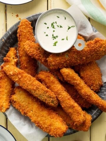 Fried pickle spears, also known as frickles, in a metal pie pan along with a cup of homemade ranch dressing.