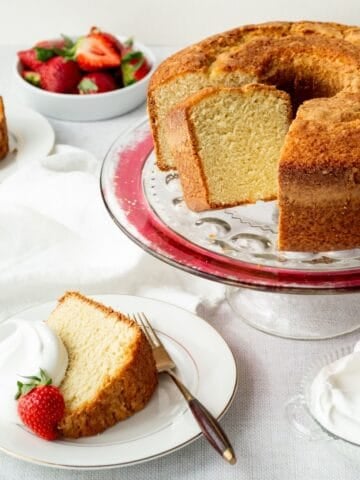 Southern sour cream pound cake on a cake stand with slices on plates topped with whipped cream and strawberries.
