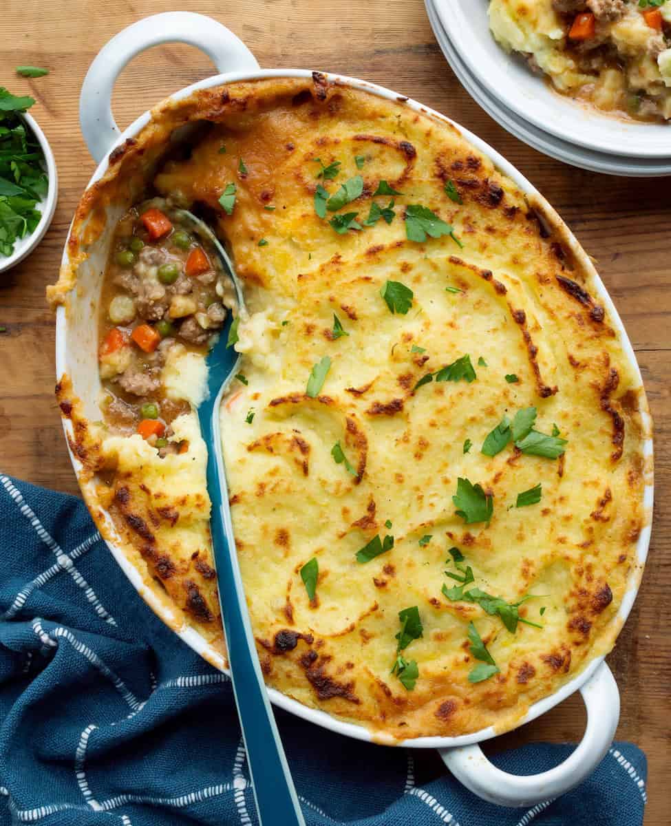 White oval casserole dish filled with scooped into shepherd's pie along with a blue spoon, blue napkin, bowl of parsley, and serving on a white plate.