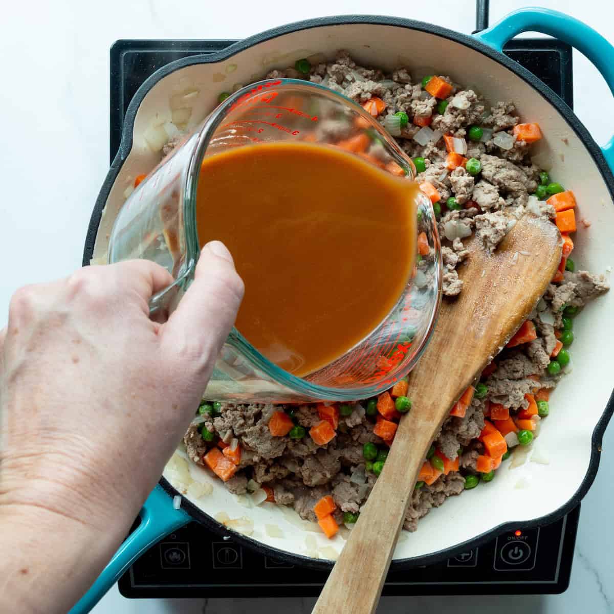 Pouring gravy ingredients into a skillet of browned turkey and frozen vegetables.