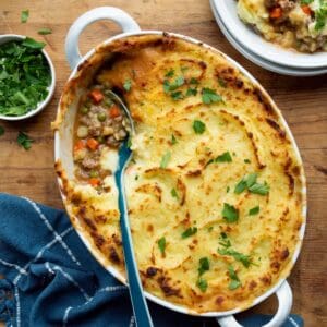 Campbell's shepherd's pie in a white oval baking dish with a blue-handled spoon, blue napkin, bowl of parsley, and a serving on a stack of white plates.
