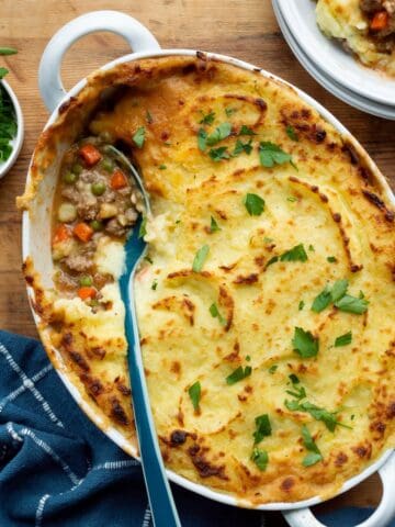 Campbell's shepherd's pie in a white oval baking dish with a blue-handled spoon, blue napkin, bowl of parsley, and a serving on a stack of white plates.