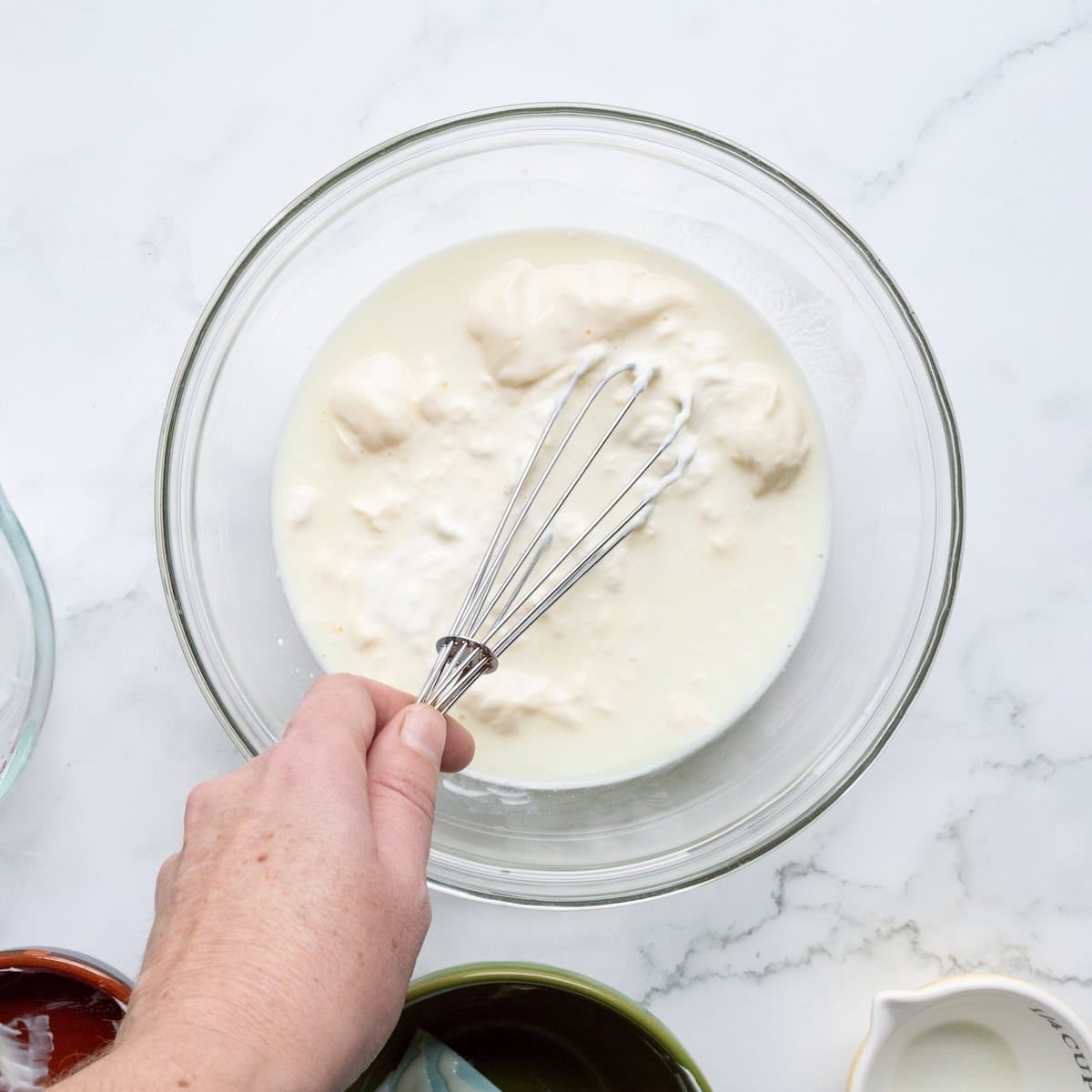 Whisking together buttermilk, mayonnaise, sour cream, and dill pickle juice in a glass bowl.