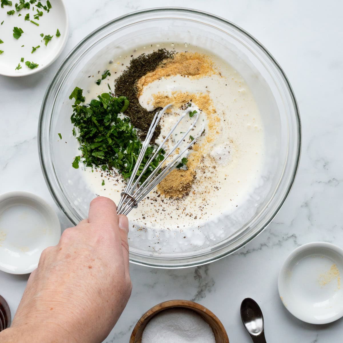 Adding seasonings and herbs to the homemade ranch dressing base in a glass bowl.