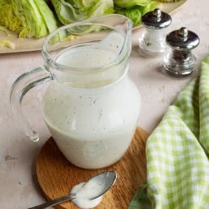 Pitcher of copycat Chili's ranch recipe on a wooden coaster with a platter of iceberg lettuce wedges in the background.